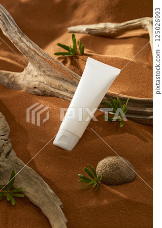 Cosmetic tube lying on brown sand with some barren trees. desert shrub growing from the sand with many lush green leaves placed around rough pebbles. Sunlight shining on objects in the dry desert. 125026993
