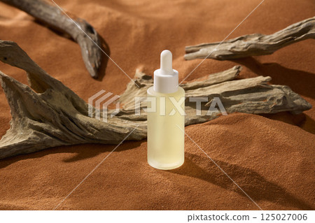 A rough, rotten tree trunk with peeling bark lies on dry brown sand. An unlabeled cosmetic dropper bottle contains a yellow essence. The sand surface is uneven with several raised areas. A rough, rotten tree trunk with peeling bark lies on dry brown sand. An unlabeled cosmetic dropper bottle contains a yellow essence. The sand surface is uneven with several raised areas. 125027006