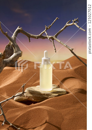 Rough rock placed in a sand pit with a serum dropper bottle displayed on top. Thorny branches grow around it. Dry and brown grain sand forms sand dunes. Tree shadows cast straight onto the sand. 125027012