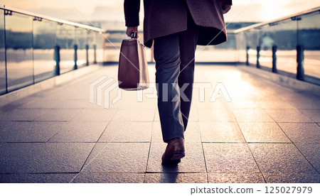 a businessman walking with a leather briefcase in hand on a modern pedestrian walkway during sunrise, symbolizing business travel, career success, leadership, and future goals, corporate, finance 125027979