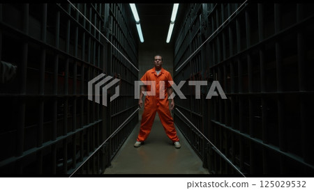 An inmate stands in a narrow prison hallway, wearing an orange jumpsuit. The strong metal bars cast shadows while bright lights illuminate the gloomy surroundings, hinting at confinement 125029532