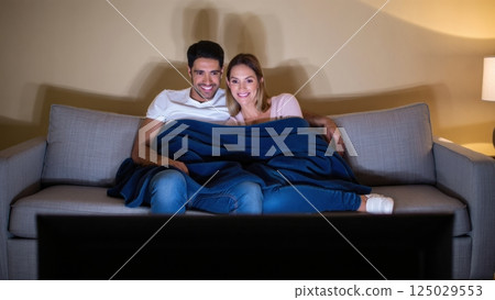 Happy couple watching tv covered by blanket on sofa 125029553