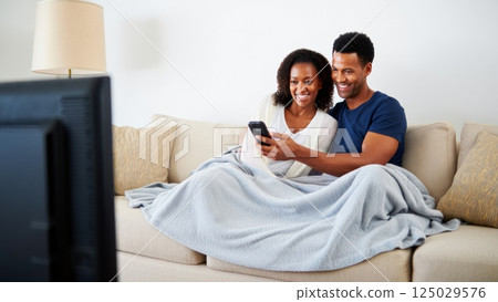 Happy couple watching tv covered by blanket on sofa Happy couple watching tv covered by blanket on sofa 125029576