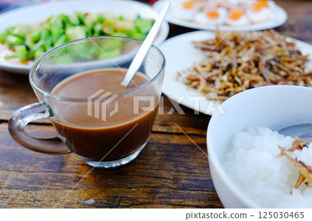 A glass cup of hot coffee with Thai food and boiled rice in white bowl on wooden table. Breakfast in the morning of Thailand A glass cup of hot coffee with Thai food and boiled rice in white bowl on wooden table. Breakfast in the morning of Thailand 125030465
