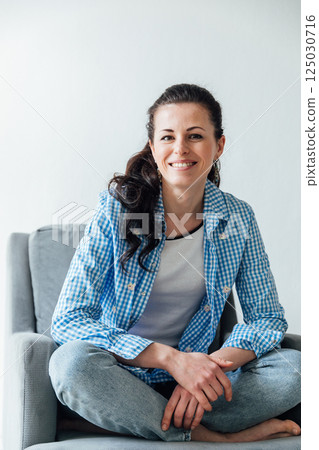 brunette woman in a blue shirt sitting on an armchair and smiling 125030716