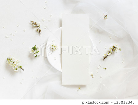 Blank card on plate near tulle and wild plums white flowers top view, romantic wedding mockup 125032343