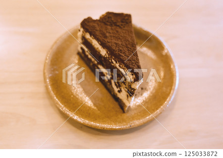 Slice of delicious tiramisu cake on ceramic plate served on wooden table in warm cozy cafe atmosphere Slice of delicious tiramisu cake on ceramic plate served on wooden table in warm cozy cafe atmosphere 125033872