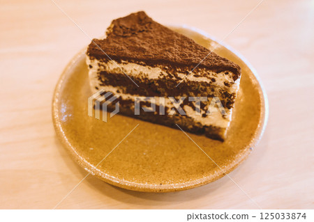Slice of delicious tiramisu cake on ceramic plate served on wooden table in warm cozy cafe atmosphere 125033874