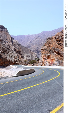 Road cuts clean line into curved desert hills, surrounded by compressed rocky walls stacked like books, glowing with layered mineral colors under midday sunlight. Canyon corridor concept. Road cuts clean line into curved desert hills, surrounded by compressed rocky walls stacked like books, glowing with layered mineral colors under midday sunlight. Canyon corridor concept. 125034082