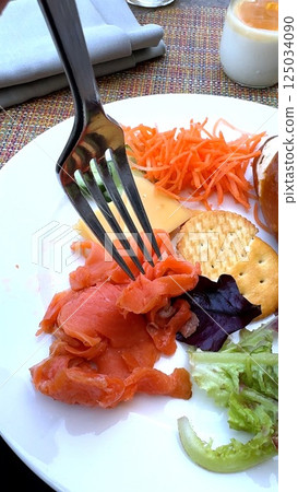 Smoked salmon curls beneath fork beside crackers, shredded carrot, crisp greens, and purple lettuce, evoking elegant health-focused brunch at scenic tourist destination escape. Smoked salmon curls beneath fork beside crackers, shredded carrot, crisp greens, and purple lettuce, evoking elegant health-focused brunch at scenic tourist destination escape. 125034090