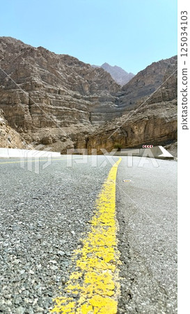 Ras Al Khaimah,UAE - March 23, 2025:Curved highway winds gracefully between canyon cliffs, road alignment flows like ribbon, sharp rocks cast strong shadows as travelers follow path into raw Ras Al Khaimah,UAE - March 23, 2025:Curved highway winds gracefully between canyon cliffs, road alignment flows like ribbon, sharp rocks cast strong shadows as travelers follow path into raw 125034103