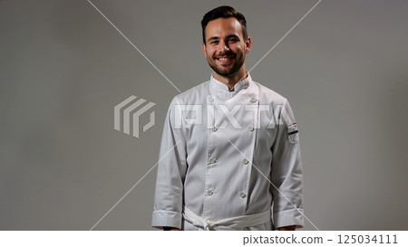 Skilled culinary professional wearing crisp white chef uniform, standing confidently with warm smile, embodying kitchen mastery against neutral gray backdrop 125034111
