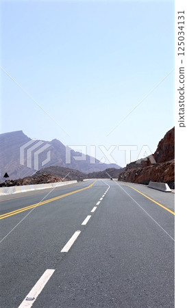 Road stretches into distant desert mountains under soft pale sky, with stone guardrails flanking the dry landscape, enhancing isolation and calm across this remote valley. Solitude escape concept. 125034131