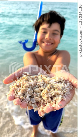 Smiling boy stretches hands full of coral fragments, ocean sparkles behind, joyful beach discovery radiates warmth and wonder, nature's raw textures bring eco learning to vibrant tourist attraction. 125034136