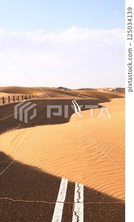 Desert road partially covered by golden sands, clear blue sky above, sand encroaches onto road suggesting timeless struggle between nature and human creation, natural dominance concept. Desert road partially covered by golden sands, clear blue sky above, sand encroaches onto road suggesting timeless struggle between nature and human creation, natural dominance concept. 125034139