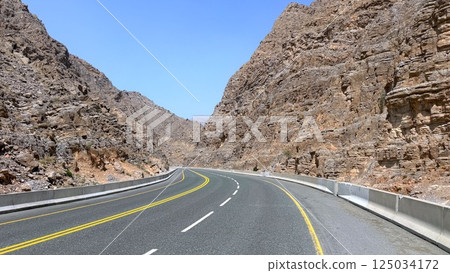 Ras Al Khaimah,UAE - March 23, 2025:Curved highway winds gracefully between canyon cliffs, road alignment flows like ribbon, sharp rocks cast strong shadows as travelers follow path into raw Ras Al Khaimah,UAE - March 23, 2025:Curved highway winds gracefully between canyon cliffs, road alignment flows like ribbon, sharp rocks cast strong shadows as travelers follow path into raw 125034172