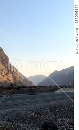 Sunlit road stretches toward distant mountains, bathed in warm golden light. Rugged peaks shaped by erosion rise against hazy sky, creating a serene landscape. Road winds through remote terrain Sunlit road stretches toward distant mountains, bathed in warm golden light. Rugged peaks shaped by erosion rise against hazy sky, creating a serene landscape. Road winds through remote terrain 125034323
