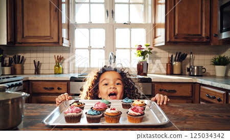 Curly haired girl smiling, holding tray of colorful cupcakes in bright kitchen, expressing cheerful culinary enthusiasm 125034342