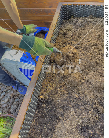 Male hands in green gloves prepare soil for planting in spring. Distributing the soil with a trowel. 125034494