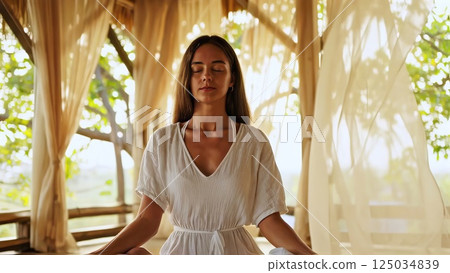 Serene female practicing mindful yoga within shaded gazebo, soft white curtains framing tranquil meditation moment in lush landscape 125034839