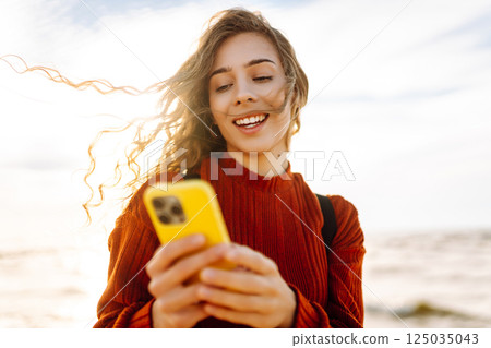 Young woman using mobile phone at beach. Cold weather. Autumn. Blogging, communication Young woman using mobile phone at beach. Cold weather. Autumn. Blogging, communication 125035043