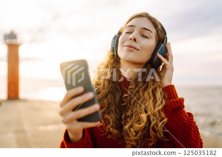Happy woman listening nice music by the waterfront while wearing headphones by a lighthouse. Happy woman listening nice music by the waterfront while wearing headphones by a lighthouse. 125035182