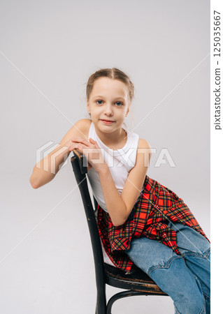 Blue-eyed preteen with light brown hair wearing white tank top, red checkered shirt and jeans, sitting posing on vintage black chair in studio with white isolated background, looking at camera. 125035667