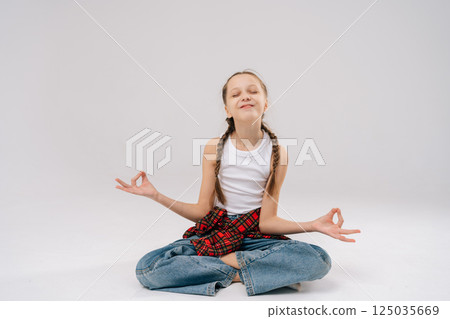 Serene child gel meditating sitting with closed eyes in lotus pose, performing mudra hand gesture in studio with white background showcasing mindfulness and tranquil inner state. 125035669