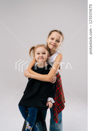 Joyful two young sisters posing for studio portrait, with older sister affectionately hugging younger sister from behind, both smiling happily looking at camera. Concept of happy sisterhood and family Joyful two young sisters posing for studio portrait, with older sister affectionately hugging younger sister from behind, both smiling happily looking at camera. Concept of happy sisterhood and family 125035670