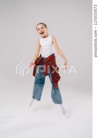 Cheerful 10 years old girl jumping mid-air with wide smile in studio with white isolated background. Happy preteen grinning widely, sporting white tank top, plaid shirt knotted at waist, baggy jeans. Cheerful 10 years old girl jumping mid-air with wide smile in studio with white isolated background. Happy preteen grinning widely, sporting white tank top, plaid shirt knotted at waist, baggy jeans. 125035673