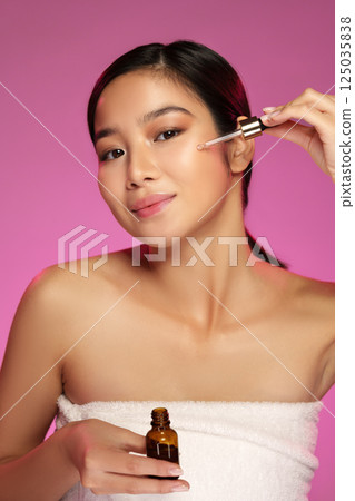 Woman applying facial serum with a dropper while smiling against a vibrant pink background 125035838
