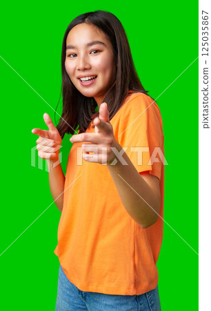 Young woman in bright orange shirt playfully poses with finger guns in front of a vibrant green background Young woman in bright orange shirt playfully poses with finger guns in front of a vibrant green background 125035867