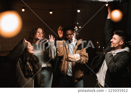 Celebration with friends as they joyfully raise their hands around a festive cake with sparklers at a cozy gathering Celebration with friends as they joyfully raise their hands around a festive cake with sparklers at a cozy gathering 125035912