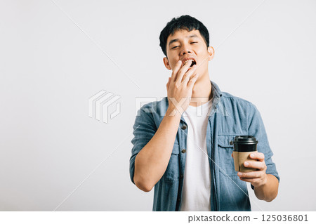 A young man, overwhelmed by fatigue, yawns with his eyes closed, coffee cup in hand. Studio shot isolated on white, his tired expression tells a relatable story. Copyspace included. 125036801
