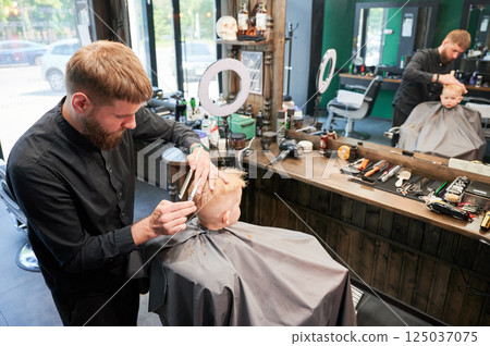 Man barber cutting little boy's hair using comb and scissors. Child getting haircut from adult male, likely barber. Professional hairdresser and cute client at modern barbershop. 125037075