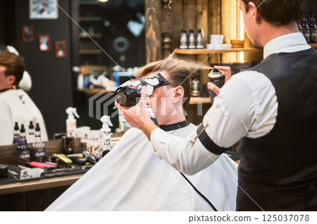 Professional hairdresser using hairdryer and hairspray for styling. Young customer getting new hairstyle. Stylish barber during process. 125037078