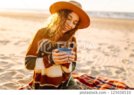 Cute woman sits on the beach drinks a hot drink from a thermos. Picnic, weekend, relax and lifestyle 125038510
