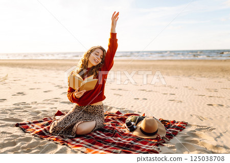 Beautiful woman sitting on blanket reading a book at beach picnic. Concept of relaxation, enjoyment. 125038708