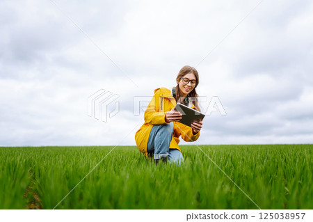 Young farmer woman in a green field, intently using a tablet. She wears a yellow jacket and glasses 125038957