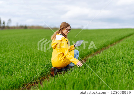 Young farmer woman in a green field, intently using a tablet. She wears a yellow jacket and glasses Young farmer woman in a green field, intently using a tablet. She wears a yellow jacket and glasses 125038959
