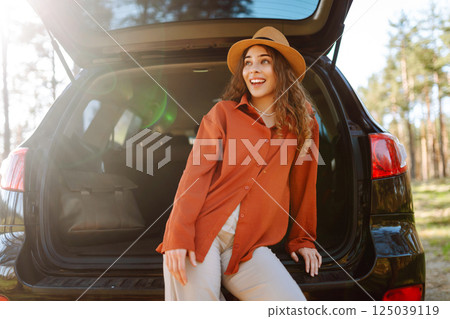 A traveler young woman smiles while sitting in the trunk of her car during a sunny day in a forest 125039119