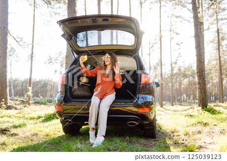 Happy woman sits on the trunk of her car and taking a selfie. Selfie while traveling by car. 125039123