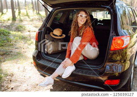 A traveler young woman smiles while sitting in the trunk of her car during a sunny day in a forest 125039127
