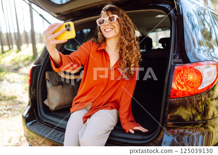Happy woman sits on the trunk of her car and taking a selfie. Selfie while traveling by car. Happy woman sits on the trunk of her car and taking a selfie. Selfie while traveling by car. 125039130