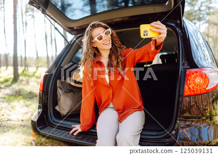 Happy woman sits on the trunk of her car and taking a selfie. Selfie while traveling by car. Happy woman sits on the trunk of her car and taking a selfie. Selfie while traveling by car. 125039131