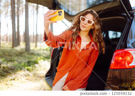 Happy woman sits on the trunk of her car and taking a selfie. Selfie while traveling by car. Happy woman sits on the trunk of her car and taking a selfie. Selfie while traveling by car. 125039132