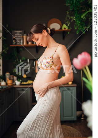 Young pregnant woman gently touching her belly in a cozy kitchen setting 125039403