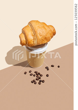 Paper cup with scattered coffee beans and fresh croissant on yellow and brown background. 125039562