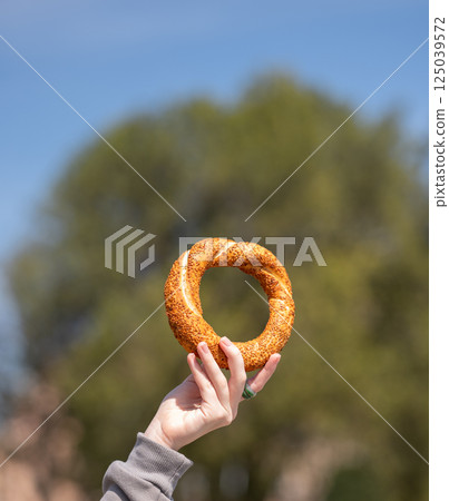A hand holds a sesame simit outdoors in front of a park of trees on a sunny day. A hand holds a sesame simit outdoors in front of a park of trees on a sunny day. 125039572