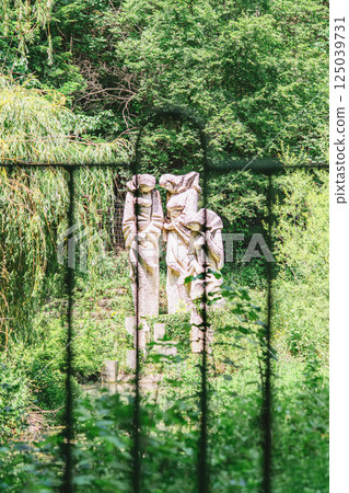 Weathered stone statues of young women figures stand hidden behind lush forest fence. Mysterious scene from folk legend, statues evokes memory, loss, and forgotten stories Weathered stone statues of young women figures stand hidden behind lush forest fence. Mysterious scene from folk legend, statues evokes memory, loss, and forgotten stories 125039731